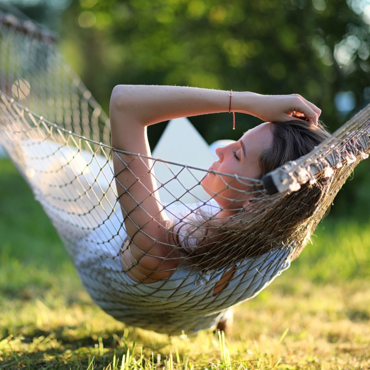 Mladá žena relaxuje v síti na letní zahradě v polostínu jako ukázka bezpečné péče o pokožku během horkých dnů.