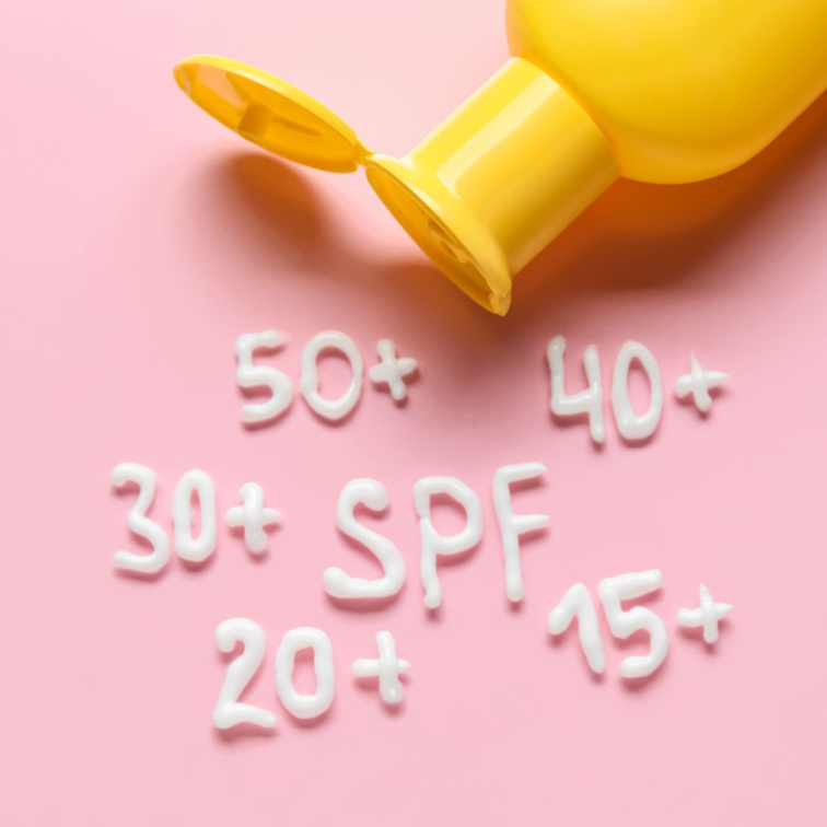 Žlutá tuba od opalovacího krému na růžovém podkladu s napsanými hodnotami SPF 15+, 20+, 30+ a 50+.
