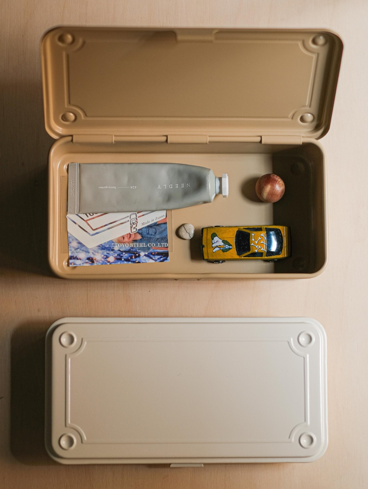 Kovový úložný box Toyo Steel T190 v béžové barvě s otevřeným víkem, uvnitř jsou drobné předměty jako tuba krému, papíry a malé objekty