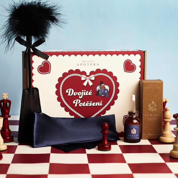 Krabička s valentínskym balíčkom Dvojité potešenie stojí na červenobielej šachovnici medzi figúrkami spolu s tmavomodrou saténovou šatkou, plácačkou s pierkom a fľaštičkou intímneho séra Venuša vedľa papierovej krabičky.