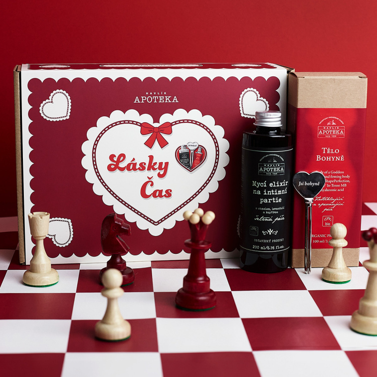 Papierová krabička valentínskeho balíčka Lásky čas stoja na červenobielej šachovnici medzi figúrkami a vedľa nej je fľaštička Umývacieho elixíru na intímne partie a krabička s krémom Telo bohyne.